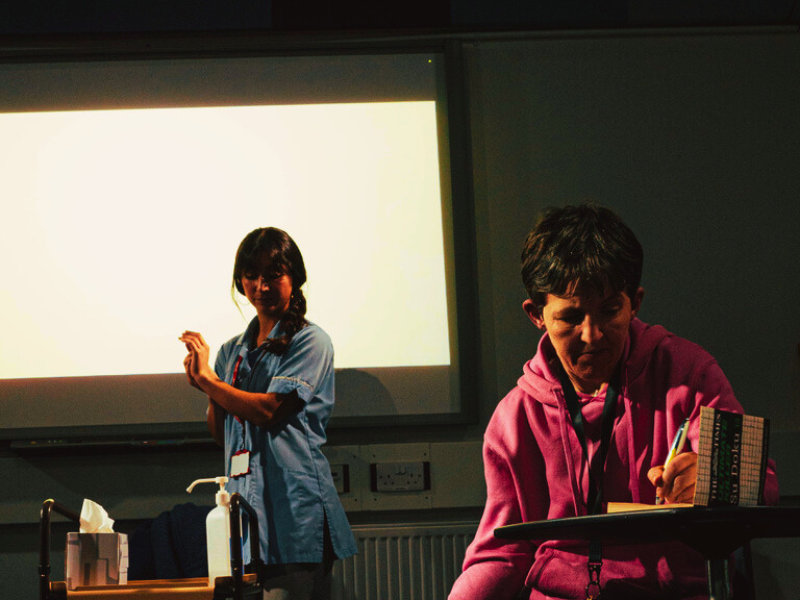 An image of a performance of Help. The performer in the foreground is sat at a table completing a sudoku. In the background a performer wears a medical tabard as they sanitise their hands.