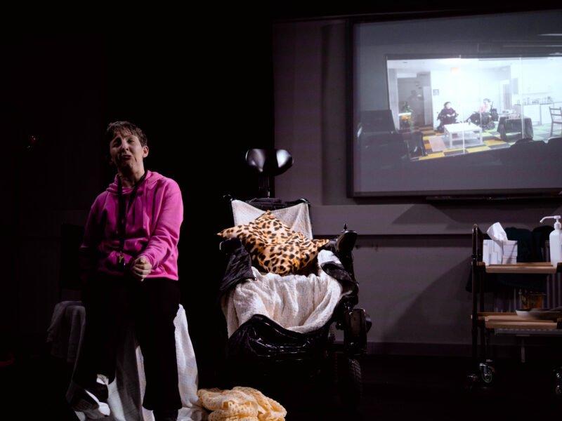 http://A%20performer%20presenting%20to%20an%20audience%20(out%20of%20shot).%20They%20are%20mid-speech%20with%20an%20image%20of%20'HELP'%20projected%20behind%20them.