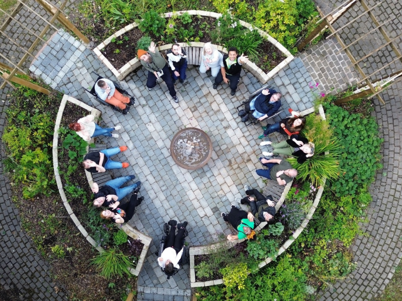 An aerial photo of DIY Theatre Company and Uncurbed Collective artists in a circle formation seated in a circular landscape in START gardens.