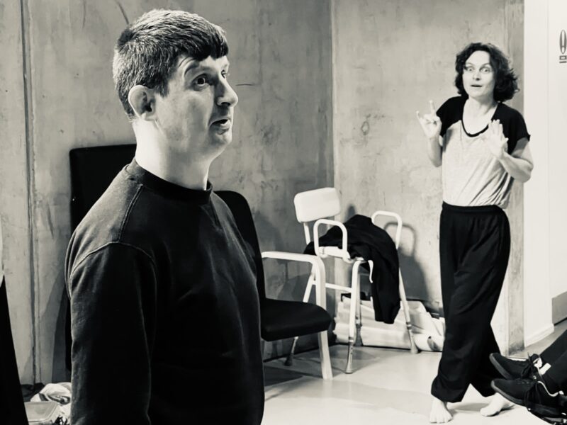 A black and white photo of Ensemble Artist Michael Gleave and Facilitator Alice Robinson. It has playful energy. In the foreground, Michael is captured in profile and he has intentional upright energy with an expression of exaggerated awe. In the background, Alice mirrors his energy and expression.